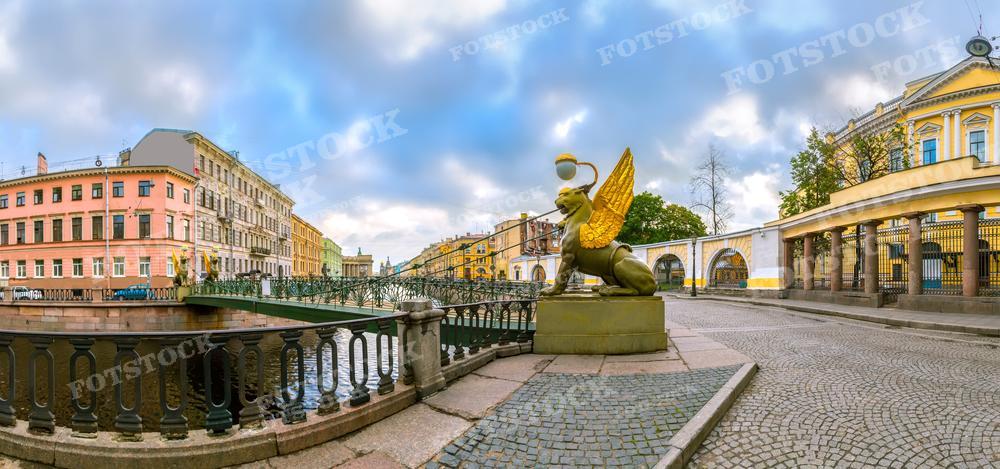 На фотографии изображен панорамный вид на один из мостов через канал в Санкт-Петербурге с золотым скульптурным изображением грифона на переднем плане. Это Банковский мост через канал Грибоедова. Погода облачная, что добавляет изображению живописности и настроения. Подобное фото может быть использовано для туристических брошюр, образовательных материалов о городе или в качестве изображения для статей о российской архитектуре и культуре.